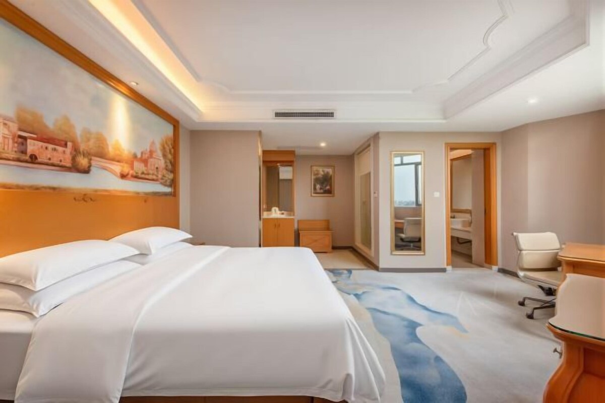 A hotel room with a large bed, a desk, and a chair. There is a painting on the wall and a view of the city.