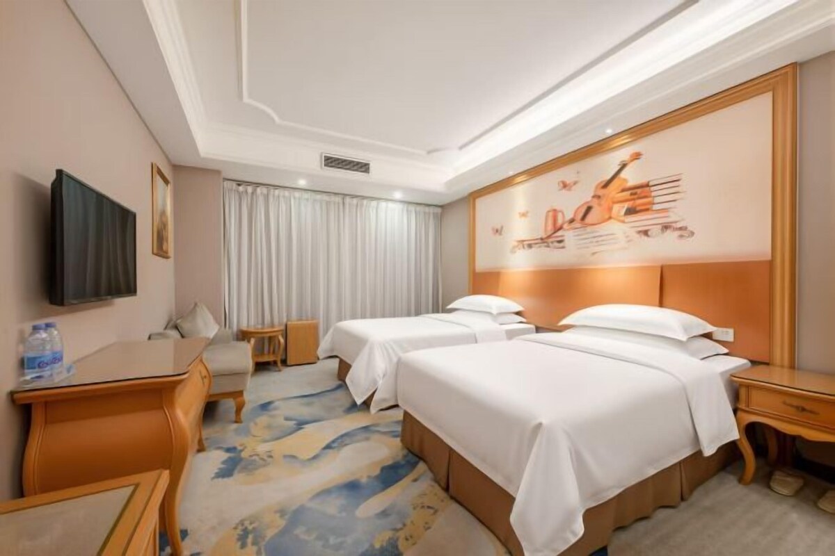 A hotel room with two beds, a desk, a chair, a television, and a large window with curtains.