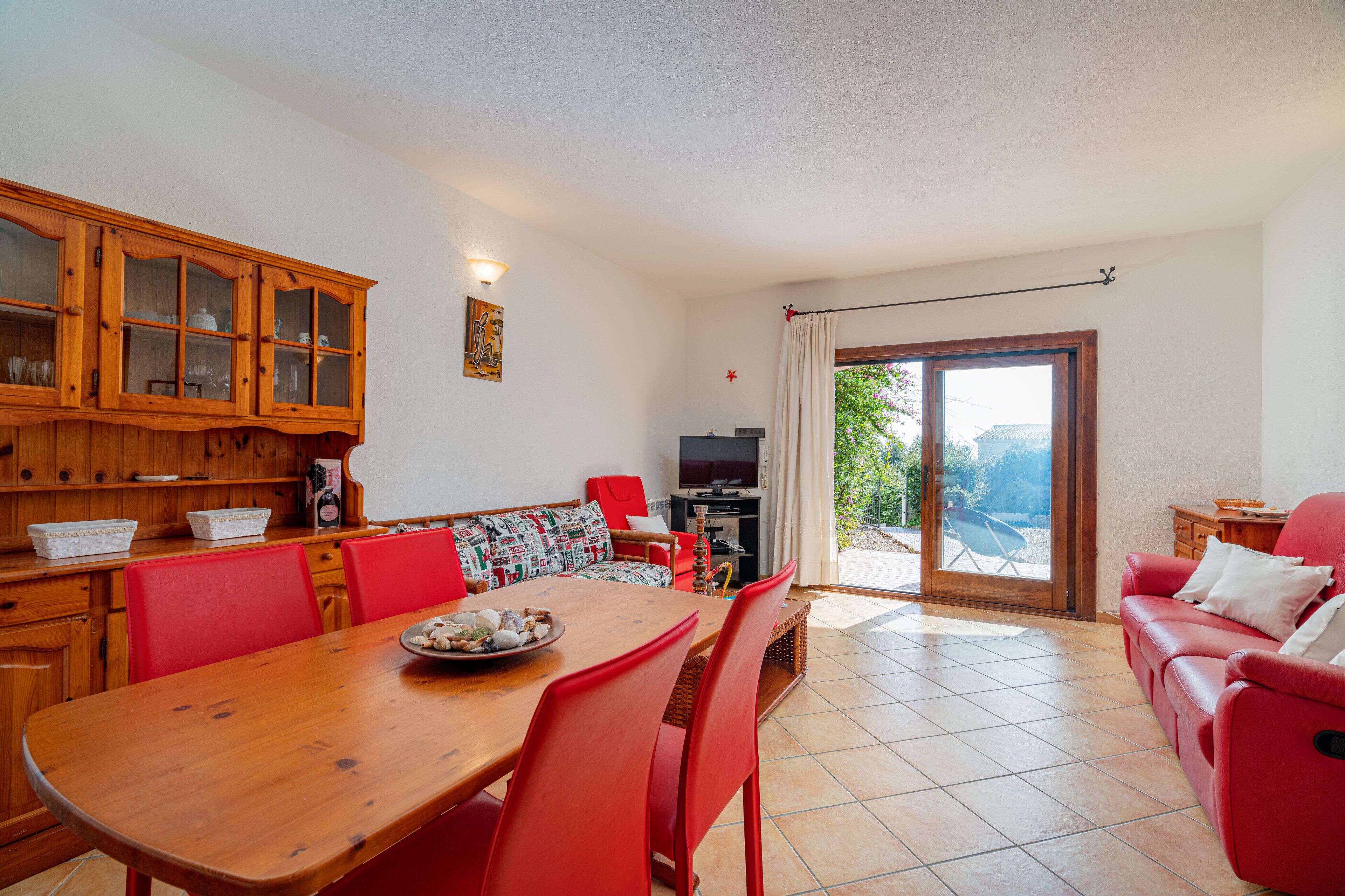 A dining area with a wooden table and red chairs, a sofa with a patterned cushion, and a television in the background.