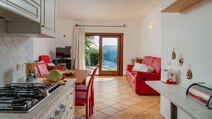 A kitchen with a stove, dining table, and chairs. There is a red sofa and a window with a view of greenery. - Casa Helen by Wonderful Italy (Olbia)