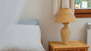 A bed with a white pillow and a beige blanket, a wicker nightstand with a lamp, and a window with a view of greenery. - Casa Helen by Wonderful Italy (Olbia)
