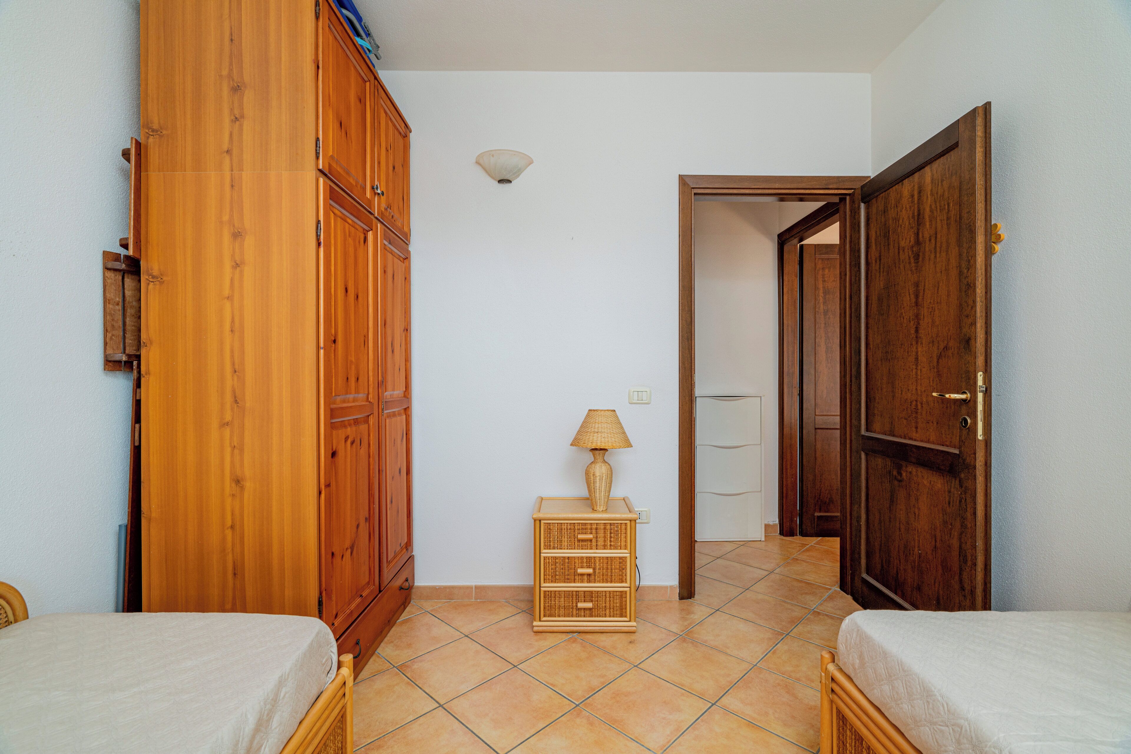A bedroom with two beds, wooden wardrobes, a small bedside table with a lamp, and a tiled floor.