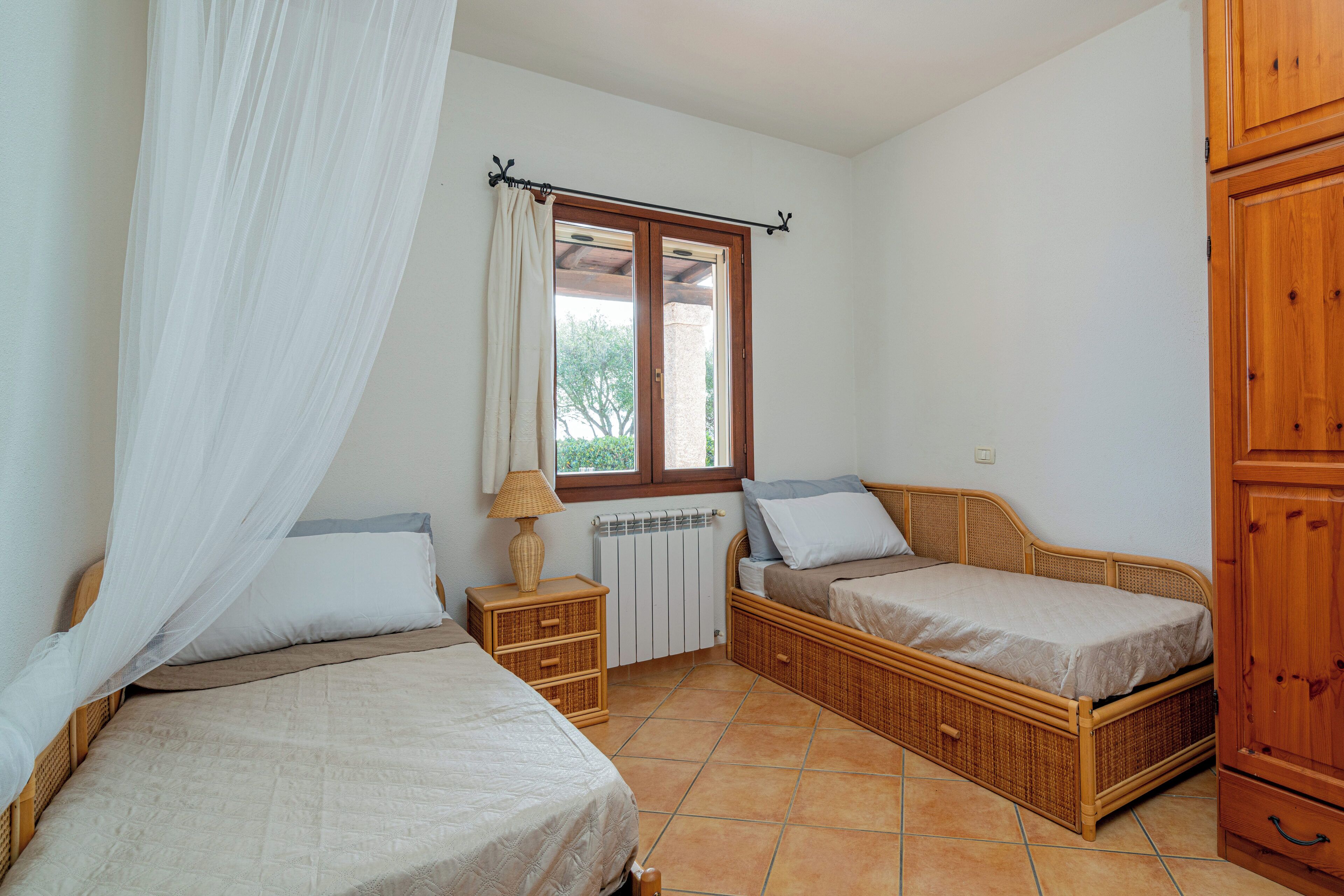 Quarto com duas camas de solteiro, guarda-roupa de madeira, mesa de cabeceira com abajur e janela com cortina.