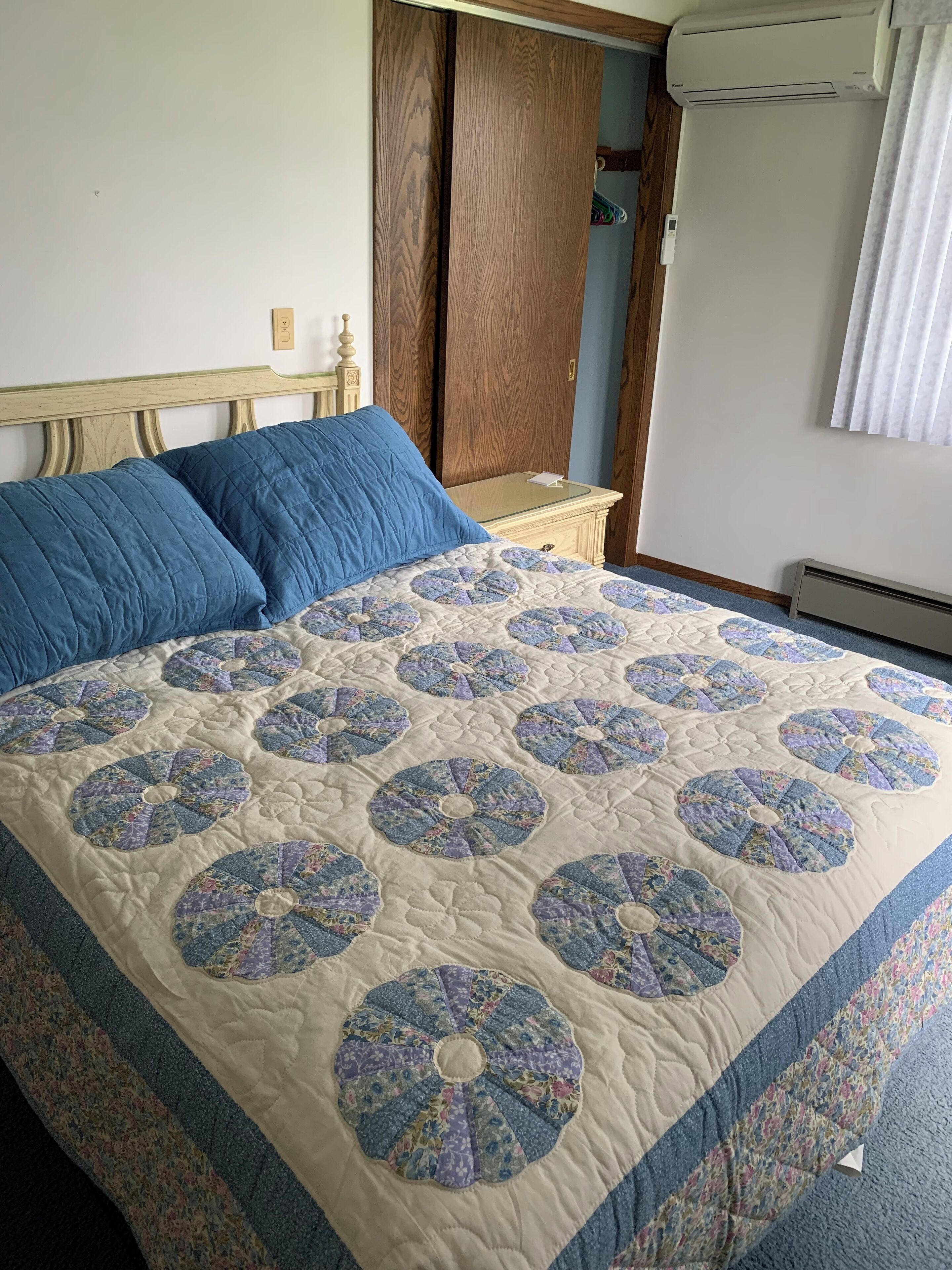 A bed with a floral quilt, a wooden wardrobe, and a white air conditioner.