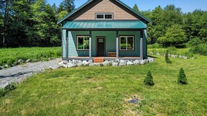 Une maison de plain-pied, à l’extérieur vert et au toit brun, entourée d’une pelouse et d’arbres.
