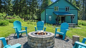 Une maison bleue avec un foyer et des fauteuils Adirondack, située dans une zone boisée.