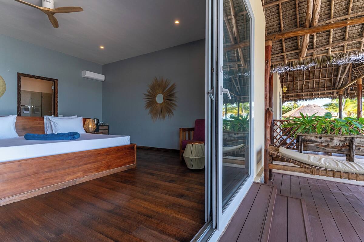 A bedroom with a wooden bed, a large mirror, and a view of a thatched-roof structure outside.