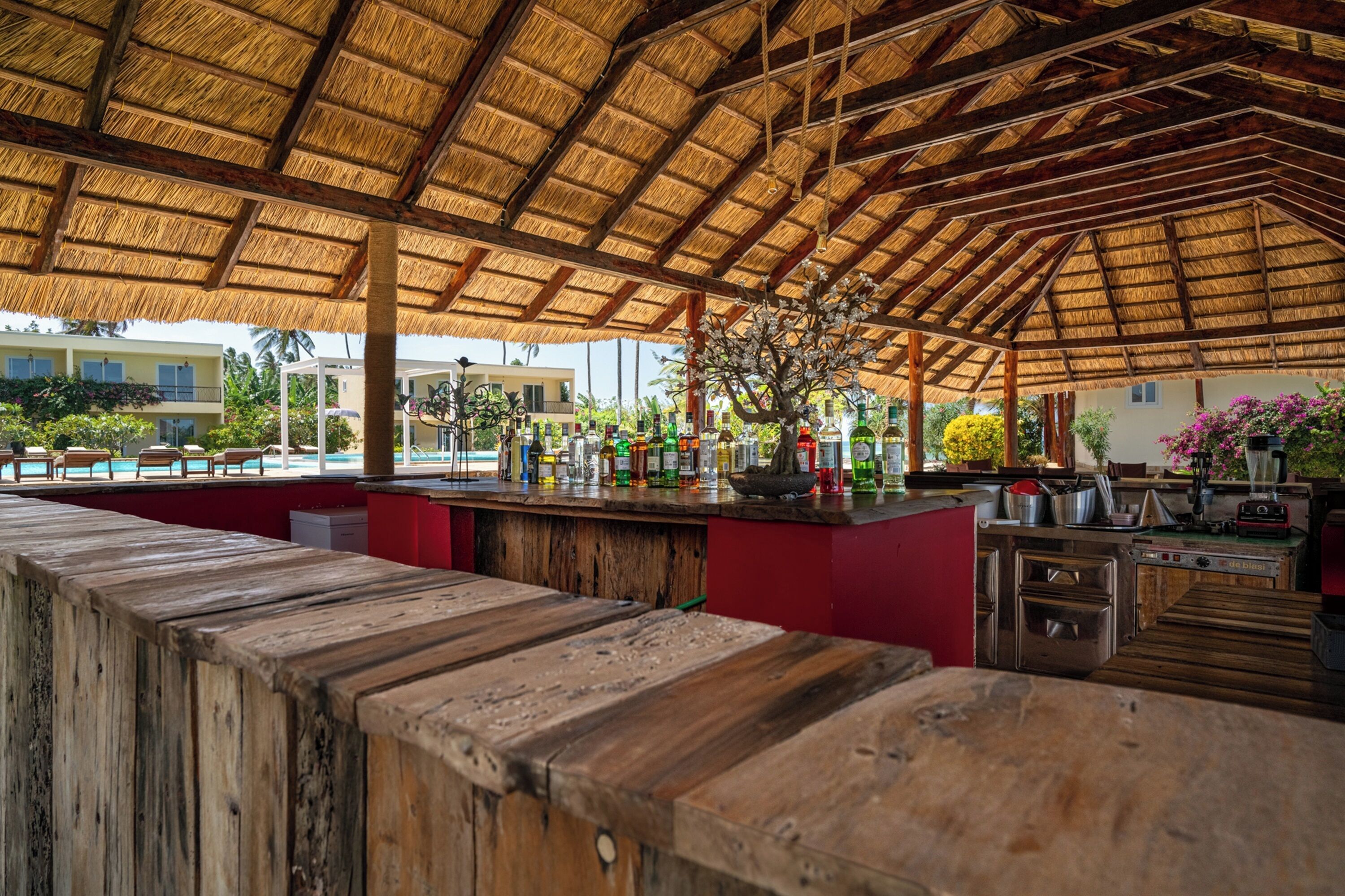 A thatched-roof outdoor bar area with a wooden counter, kitchen appliances, and a view of the surrounding buildings.