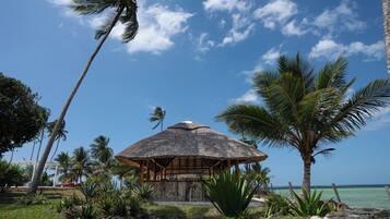 Una cabaña con techo de paja y estructura de madera, rodeada de vegetación tropical y palmeras, con vistas a un mar azul cristalino.