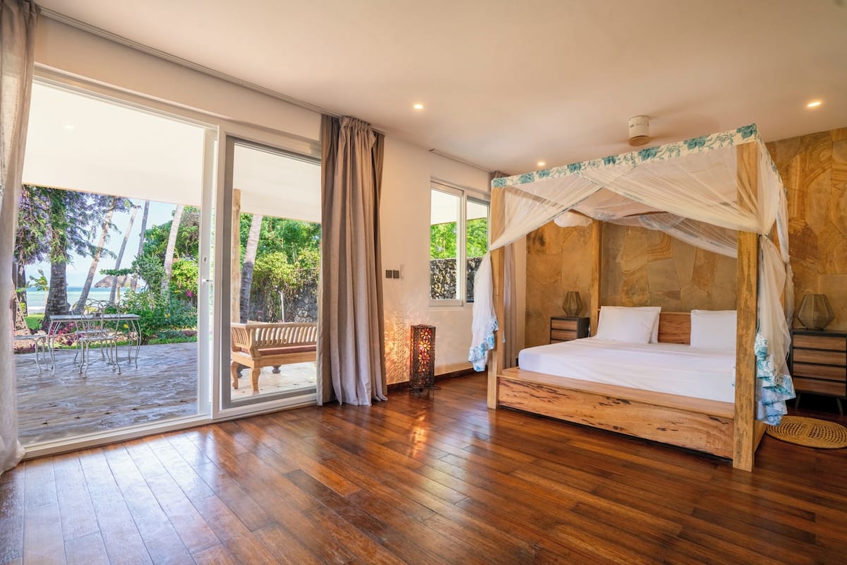 A spacious bedroom with a wooden floor, a large bed with a canopy, and a sliding glass door leading to an outdoor area with a bench and greenery.
