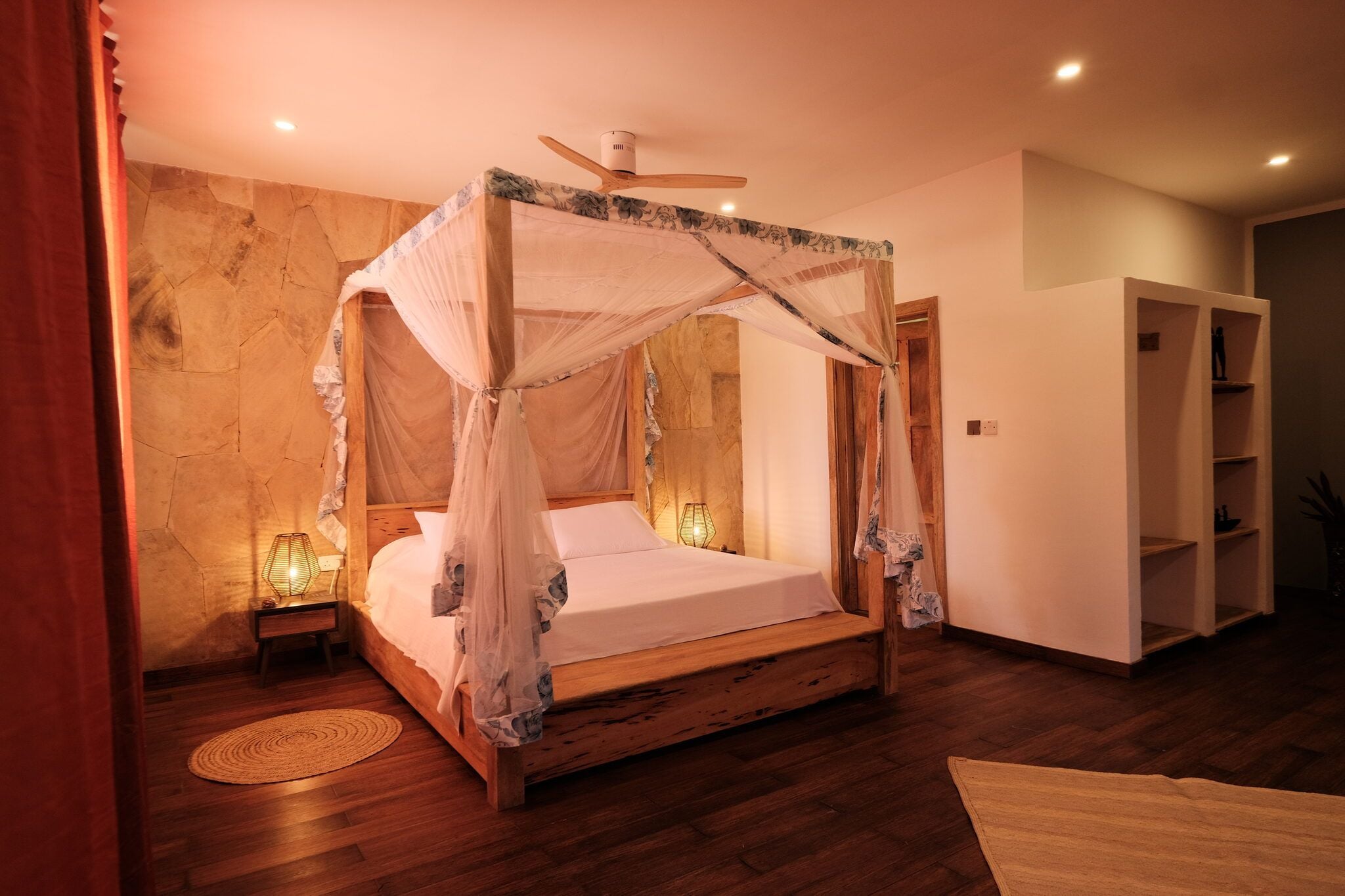 Un dormitorio con una cama con dosel, una mesita de noche con lámpara, suelo de madera y una estantería empotrada.