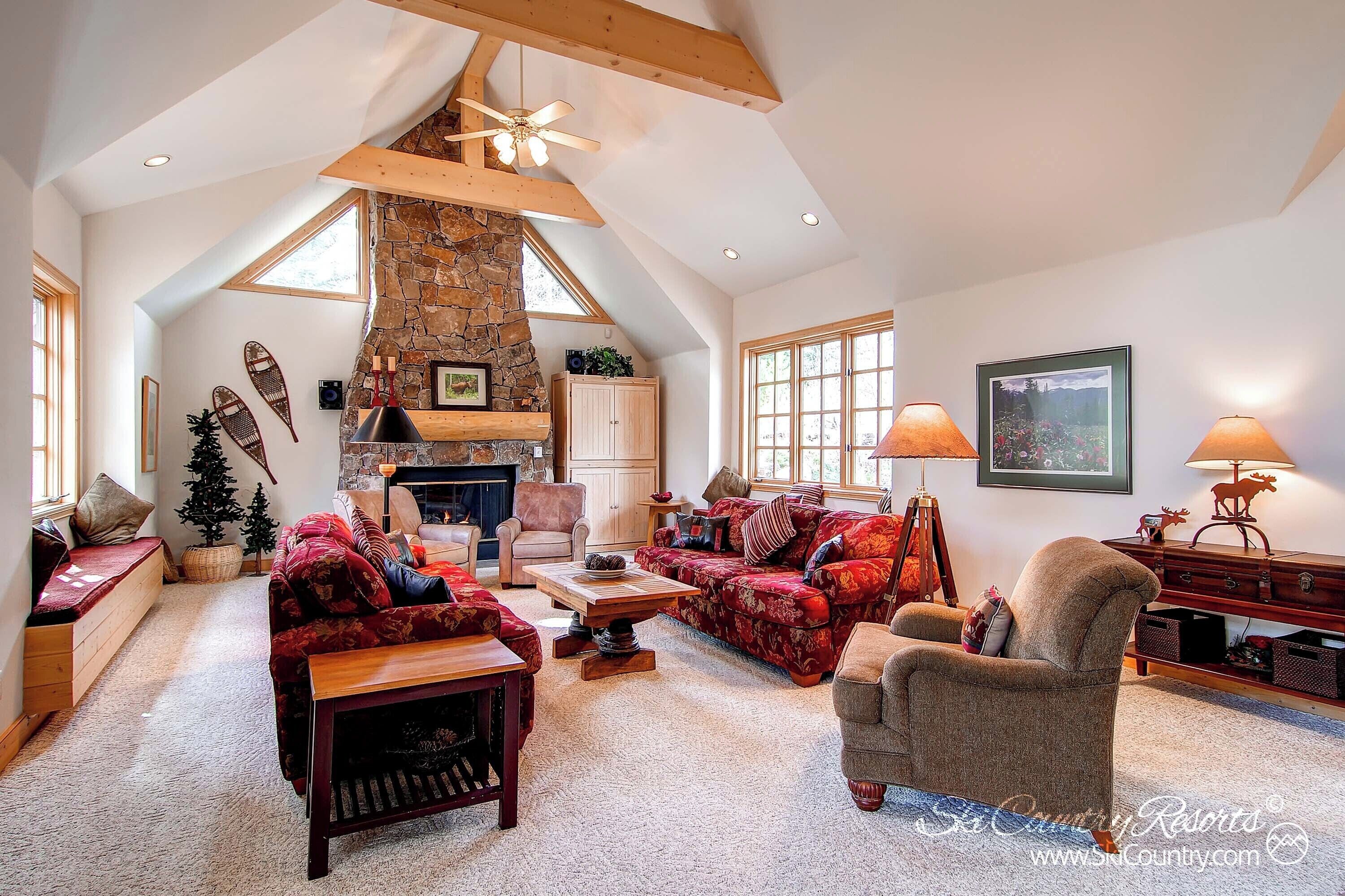 Un vaste salon doté d’un haut plafond, de poutres en bois, d’une cheminée en pierre et d’un coin salon confortable avec des meubles recouverts de tissu rouge.