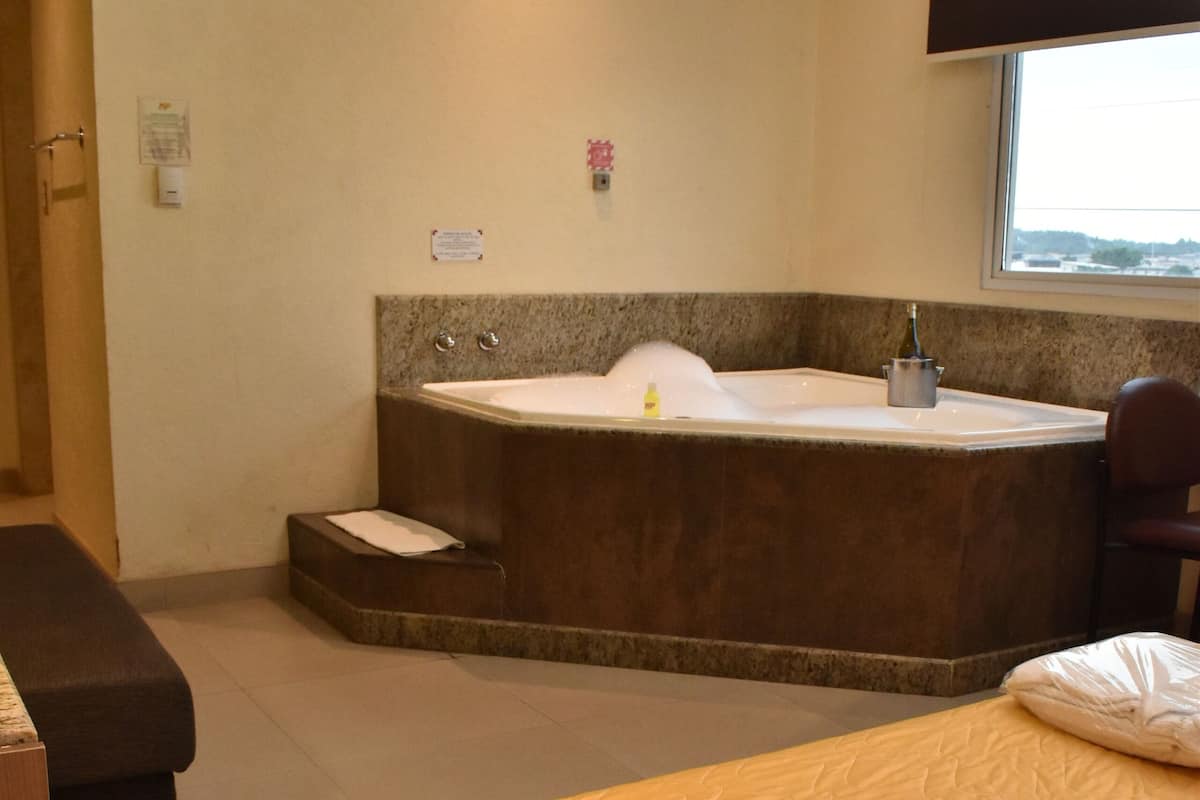 A room with a large bathtub, a bed with an orange cover, and a window with a view.
