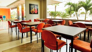 A spacious dining area with multiple tables and orange chairs, large windows with a view of palm trees, and a colorful abstract painting on the wall. - NP Hotel & Suites (Guayaquil)