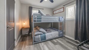 Een slaapkamer met stapelbed, plafondventilator, nachtkastje met lamp, raam met gordijnen en een motiverende quote op de muur.