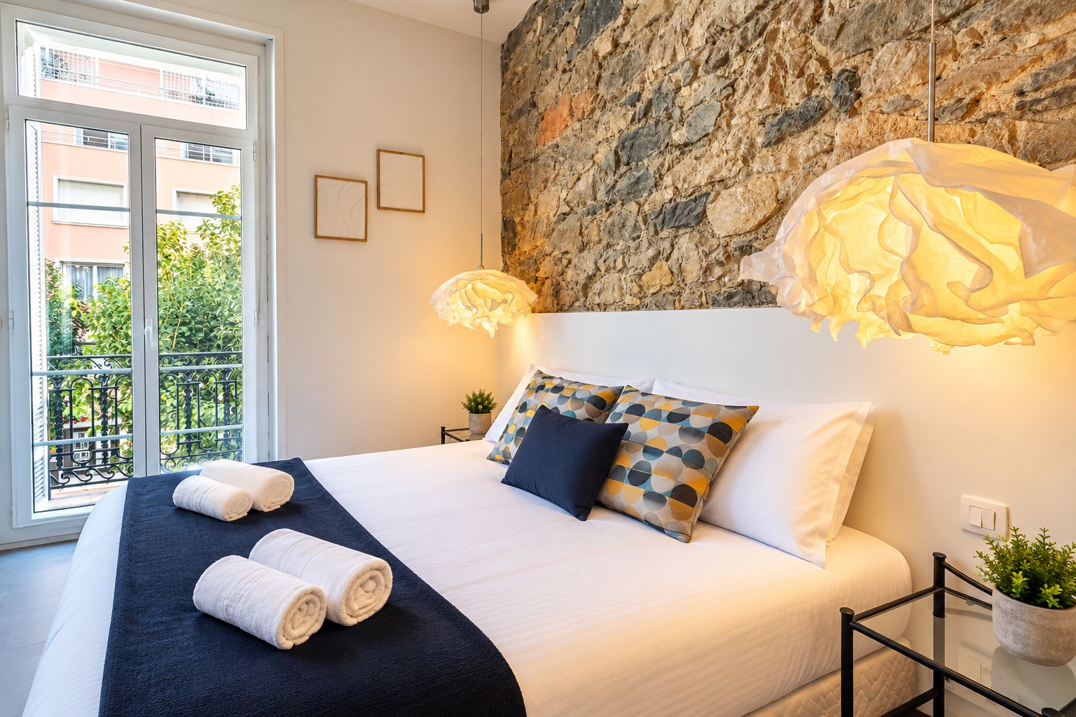 Een moderne slaapkamer met een stenen muur, een groot bed met veel kussens, een nachtkastje, een potplant en uitzicht op een balkon met groen.