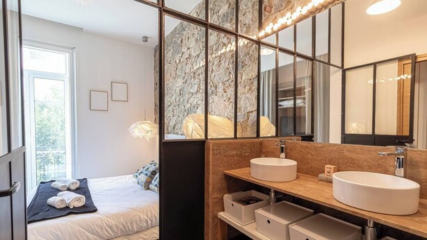 A modern bedroom with a large bed, a bedside table, and a bathroom with a large mirror, a sink, and storage drawers. - Elegant Belle Époque Apartment in Central Menton (Menton)