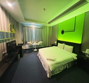 Une chambre d’hôtel avec un lit, un bureau avec une télévision, une chaise, une petite table avec une lampe et un mur d’accent vert.