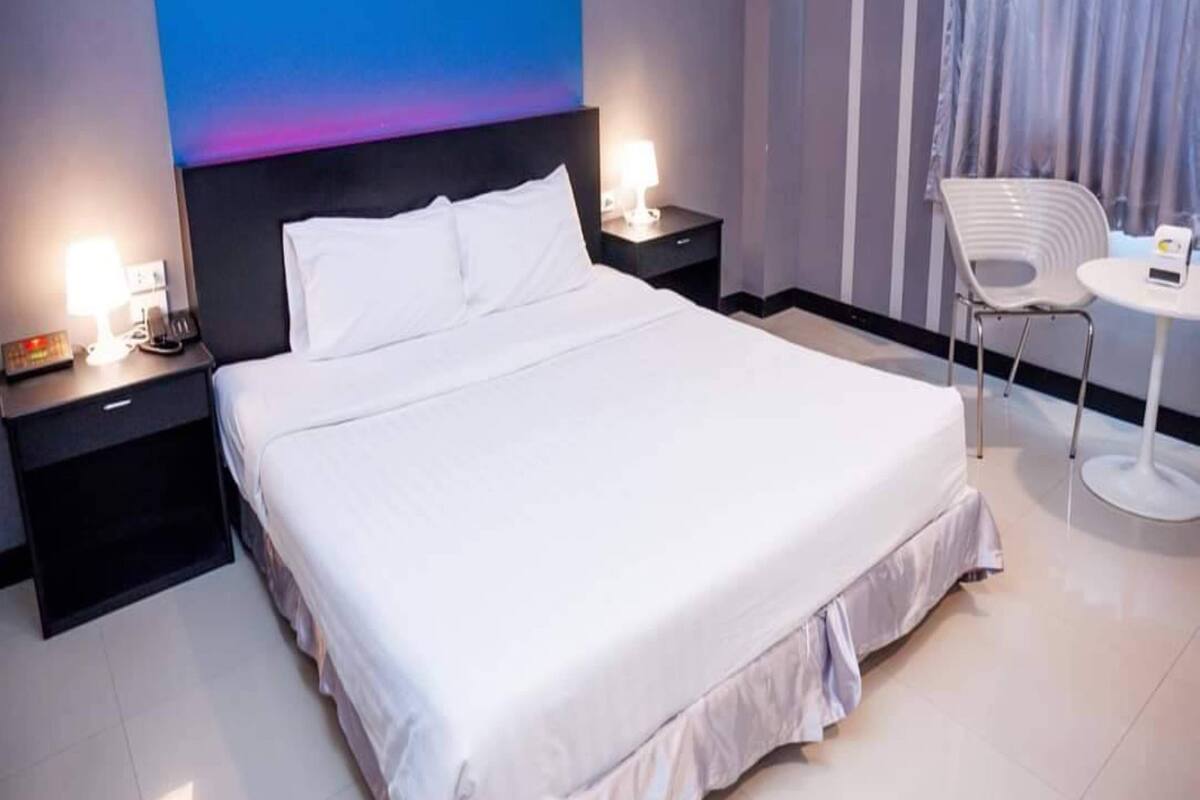 A modern hotel room with a large bed, a nightstand with a lamp, a small table with a chair, and a wall with a geometric design.