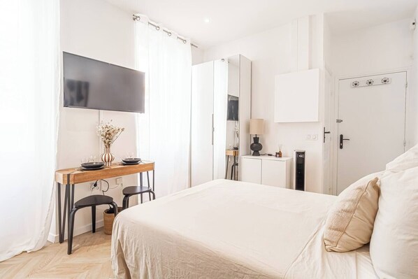A modern hotel room with a large bed, a desk with a vase of flowers, a flat-screen TV, and a wardrobe.