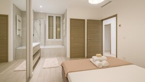 A modern bedroom with a large bed, a walk-in closet, a bathroom with a shower, and a window with shutters.