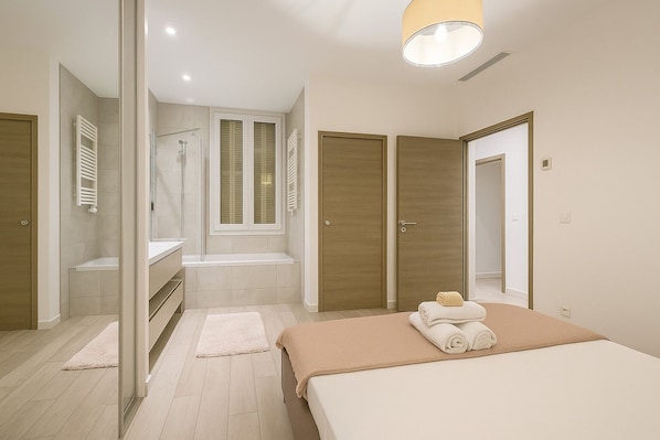 A modern bedroom with a large bed, a walk-in closet, a bathroom with a shower, and a window with shutters.