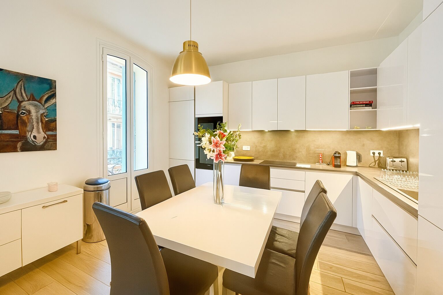A modern kitchen with white cabinets, a dining table with chairs, a pendant light, and a vase with flowers.