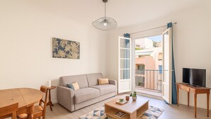 En stue med sofa, sofabord og TV-møbel, et spisebord med stole, samt et stort vindue med udsigt til bygninger.