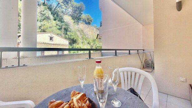 A balcony with a table set for two, featuring croissants, a bottle of orange juice, and two wine glasses. - 1 bedroom apartment • Near Monaco Train Station (Beausoleil)