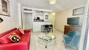 A living room with a red sofa, a glass coffee table, a blue armchair, a TV, and a kitchen in the background. - 1 bedroom apartment • Near Monaco Train Station (Beausoleil)