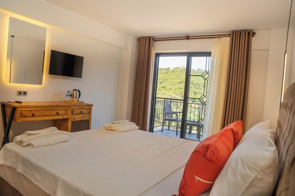 A hotel room with a bed, a wooden desk with a flat-screen TV, a balcony with a chair, and a view of greenery.