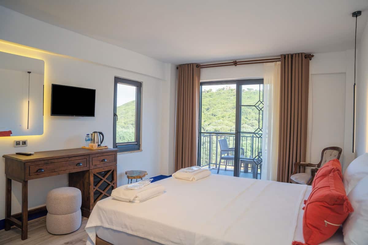 A hotel room with a bed, a desk, a TV, and a balcony with a view of greenery.