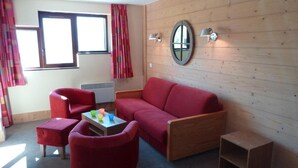 Une pièce chaleureuse avec un canapé rouge, un fauteuil assorti, une petite table en bois et un pouf rouge. On y trouve une fenêtre avec des rideaux, une lampe fixée au mur et un miroir.