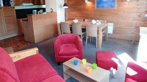 Un salon chaleureux avec des canapés rouges, une table à manger en bois et une cuisine en arrière-plan.