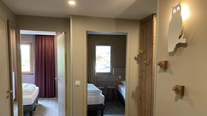 A hotel room with a wooden floor, two beds, a window with curtains, and a wooden door.