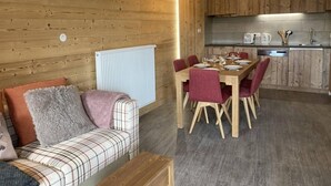 A cozy room with a wooden wall, a sofa with plaid cushions, a dining table with red chairs, and a kitchen area with wooden cabinets and a countertop.