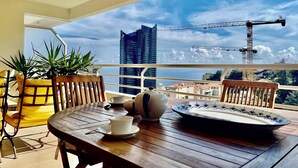 A balcony with a wooden table, chairs, and potted plants. A cityscape with buildings and cranes is visible in the background. - 2 rooms in luxury residence bordering Monaco (Beausoleil)