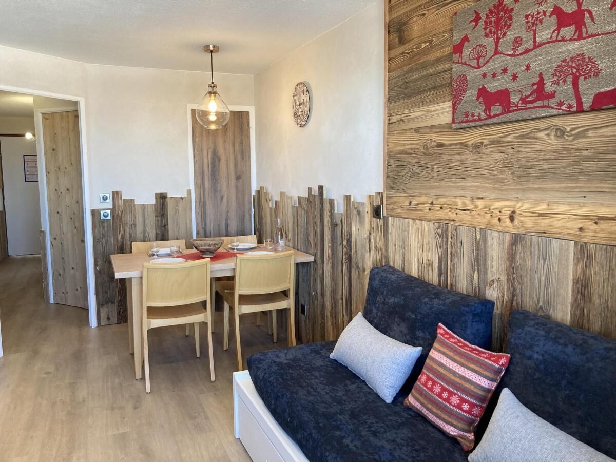 Um quarto aconchegante com um sofá azul escuro, duas cadeiras de madeira, uma mesa de jantar e uma parede de destaque em madeira com uma obra de arte estampada em vermelho e branco.