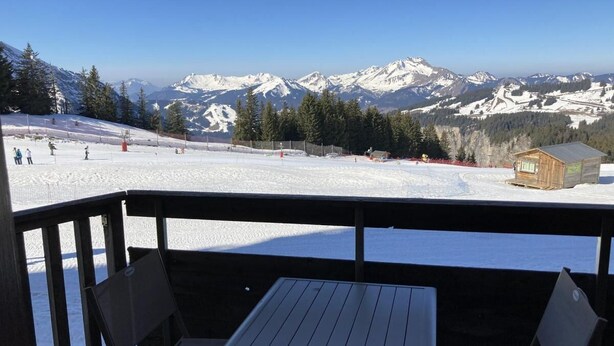 Eine Holzdielen-Terrasse mit Tisch und Stühlen mit Blick auf eine verschneite Berglandschaft, in der Ferne ein kleines Holzhaus.
