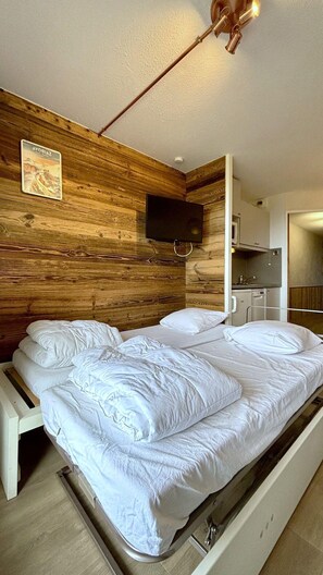 A bedroom with a wooden headboard, a bed with white bedding, a television mounted on the wall, and a kitchenette area.