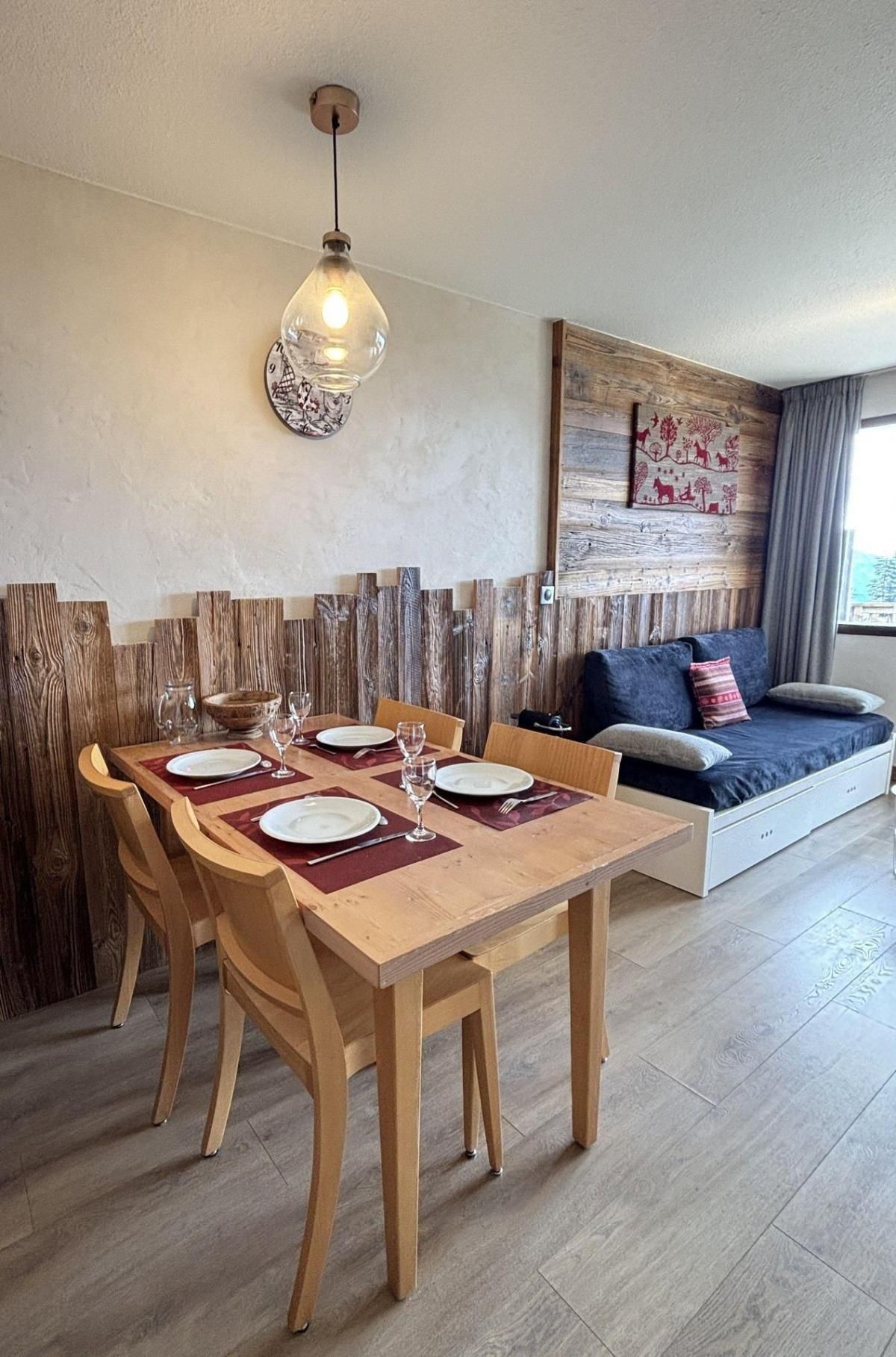 Un comedor con una mesa de madera dispuesta para cuatro personas, una lámpara colgante y una pared rústica con paneles de madera.