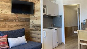 Ein kompakter Wohnraum mit Sofa, einem Flachbildfernseher an einer Holzwand, einer Küchenzeile mit Mikrowelle und einer kleinen Essecke.