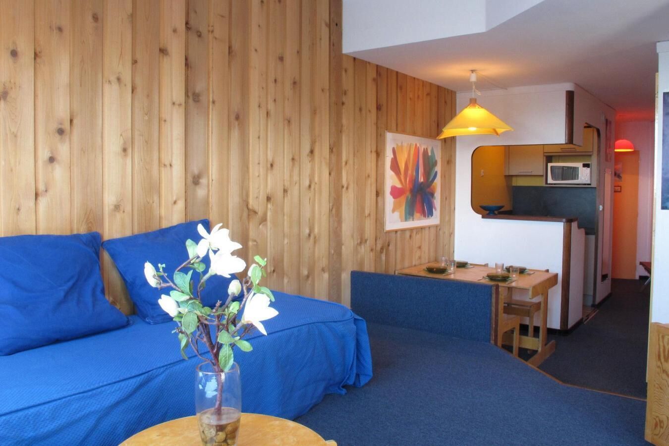 A cozy room with a blue sofa, a small table with a vase of flowers, and a kitchenette in the background.
