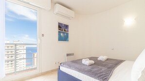 A bedroom with a bed, a window with a view of buildings and water, and an air conditioning unit. - Beautiful apartment near Monaco (Beausoleil)