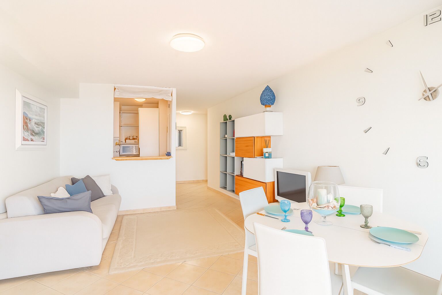 A modern, well-lit living room with a white sofa, a dining table with chairs, and a kitchen area in the background.