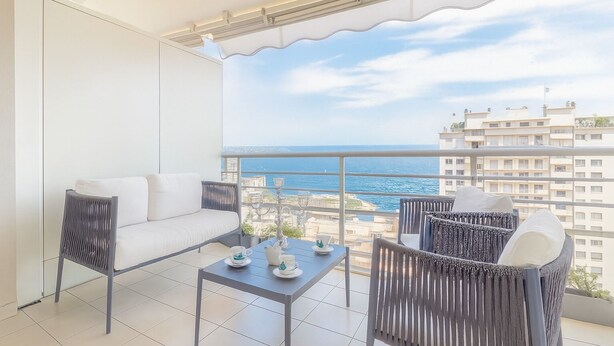 A balcony with a sofa and chairs, a table with cups and a teapot, and a view of the sea and buildings. - Beautiful apartment near Monaco (Beausoleil)