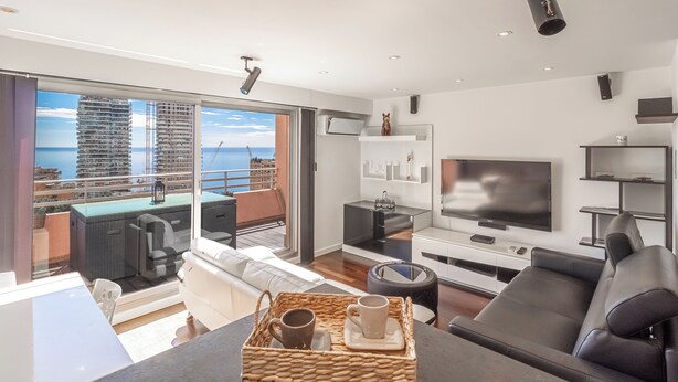 A modern living room with a large TV, comfortable seating, a dining area, and a balcony with a city view.