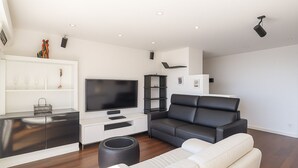 A modern living room with a flat-screen TV, a black leather sofa, a white leather couch, and a small round ottoman.
