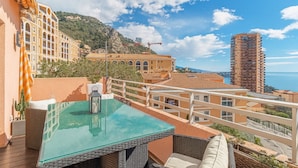 A rooftop terrace with a glass table, wicker furniture, and a view of the sea and surrounding buildings.
