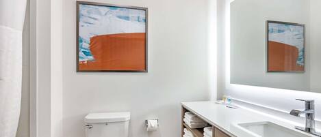 A modern bathroom with a toilet, a sink, and a large mirror. There are towels on a shelf and a framed abstract painting on the wall.